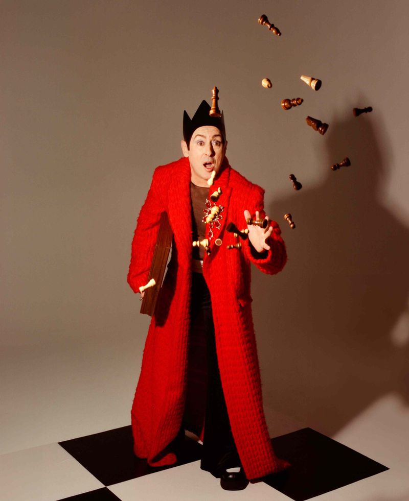 Full shot of a man in a red coat and crown juggling chess pieces in a studio setting with a black-and-white checkered floor.