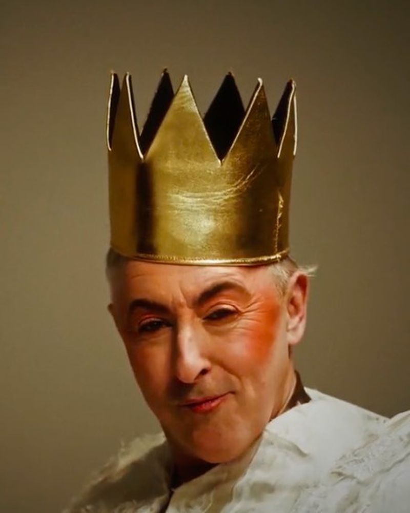 Man in gold crown with pale face makeup looking directly at camera indoors in portrait. Head and shoulders.