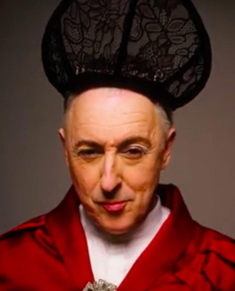 Close-up portrait of an adult male with a black lace hat and a red robe looking at the camera against a neutral backdrop.