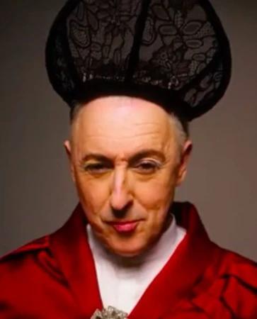 Close-up portrait of an adult male with a black lace hat and a red robe looking at the camera against a neutral backdrop.