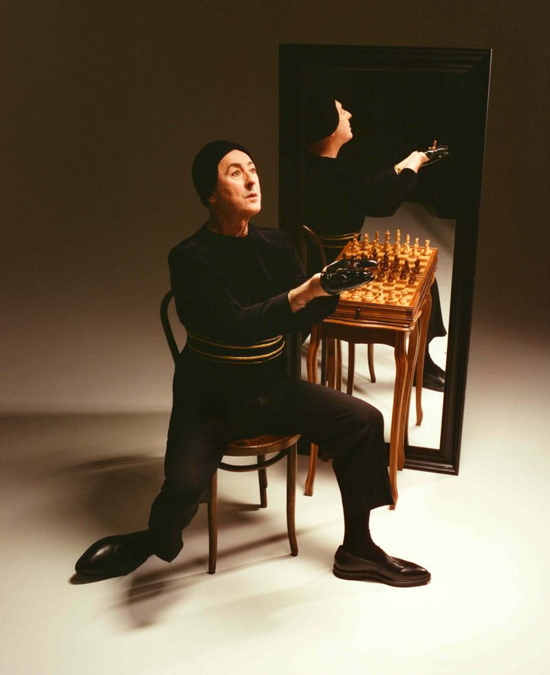Man in dark clothing seated at a table with a chess set, reflected in a mirror, with the same figure looking off into the distance.