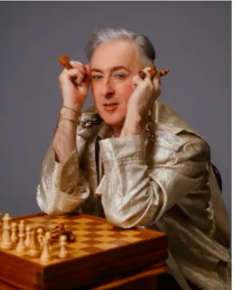 Adult male in a shiny gold jacket sits indoors at a chess board holding chess pieces up to his eyes in a unique studio portrait.