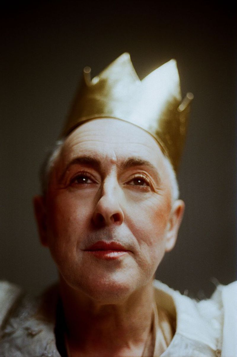 Headshot of an adult man wearing a gold crown, wearing theatrical makeup and white frilled top, looking up against a dark background.