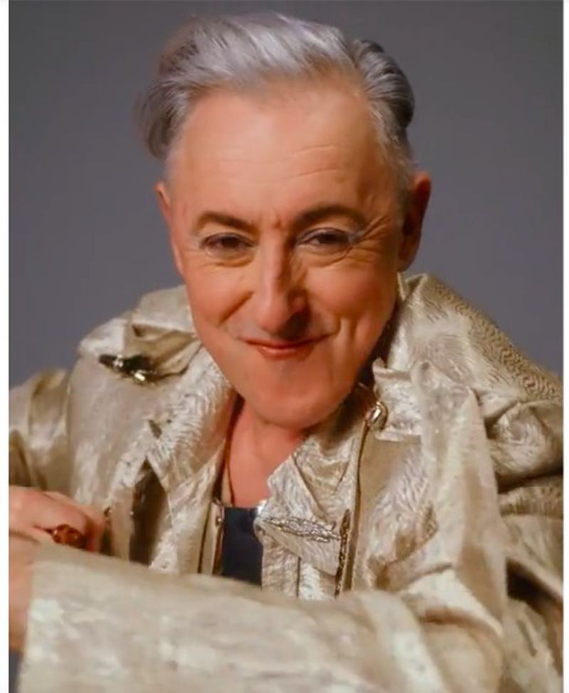Close-up studio portrait of a smiling adult with gray hair, wearing a gold jacket with an open collar, set against a solid gray backdrop.