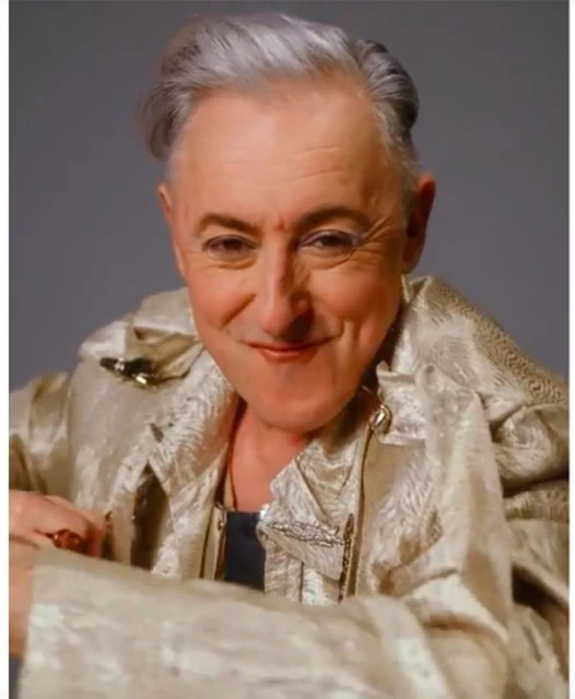 Close-up studio portrait of a smiling adult with gray hair, wearing a gold jacket with an open collar, set against a solid gray backdrop.