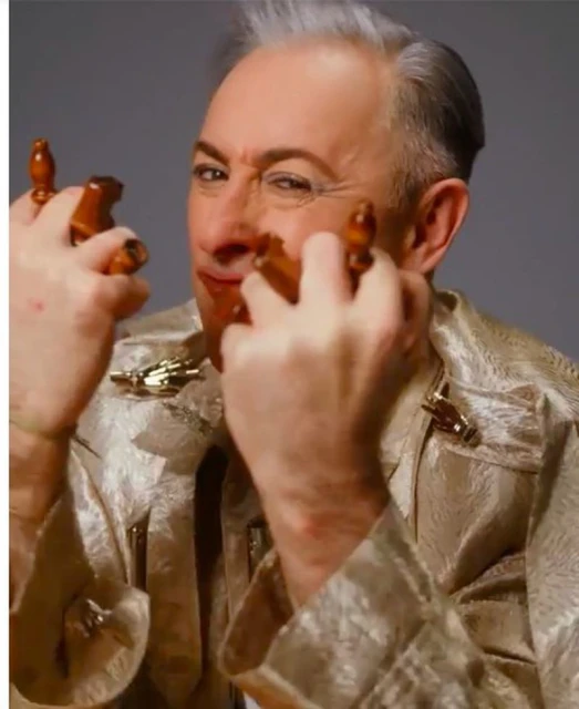 Close-up of a mature adult male in a patterned shirt, holding wood sculptures, set against a neutral background, creating a distinctive portrait.