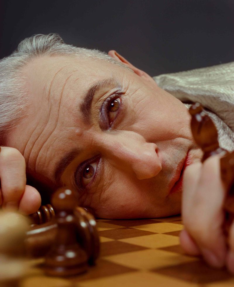 Close-up portrait of mature adult male with grey hair, wearing makeup, looking down at chess board and pieces in a photo studio.