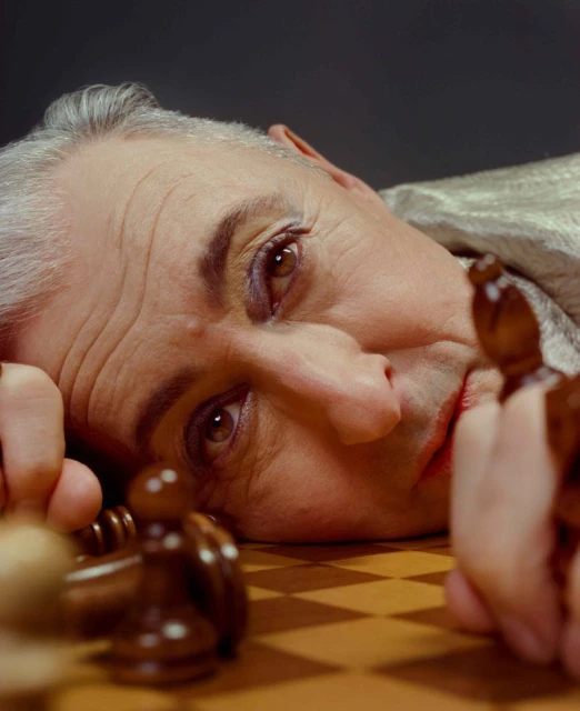 Close-up portrait of mature adult male with grey hair, wearing makeup, looking down at chess board and pieces in a photo studio.