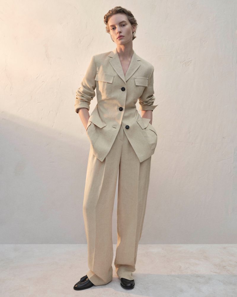 Woman in a beige pantsuit poses in front of a neutral wall. The outfit features a blazer with black buttons and relaxed-fit trousers.