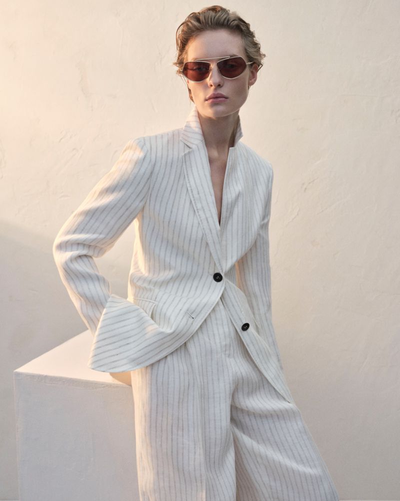 Woman in a striped linen pantsuit and brown sunglasses poses against a cream-colored wall and geometric form in soft studio lighting.