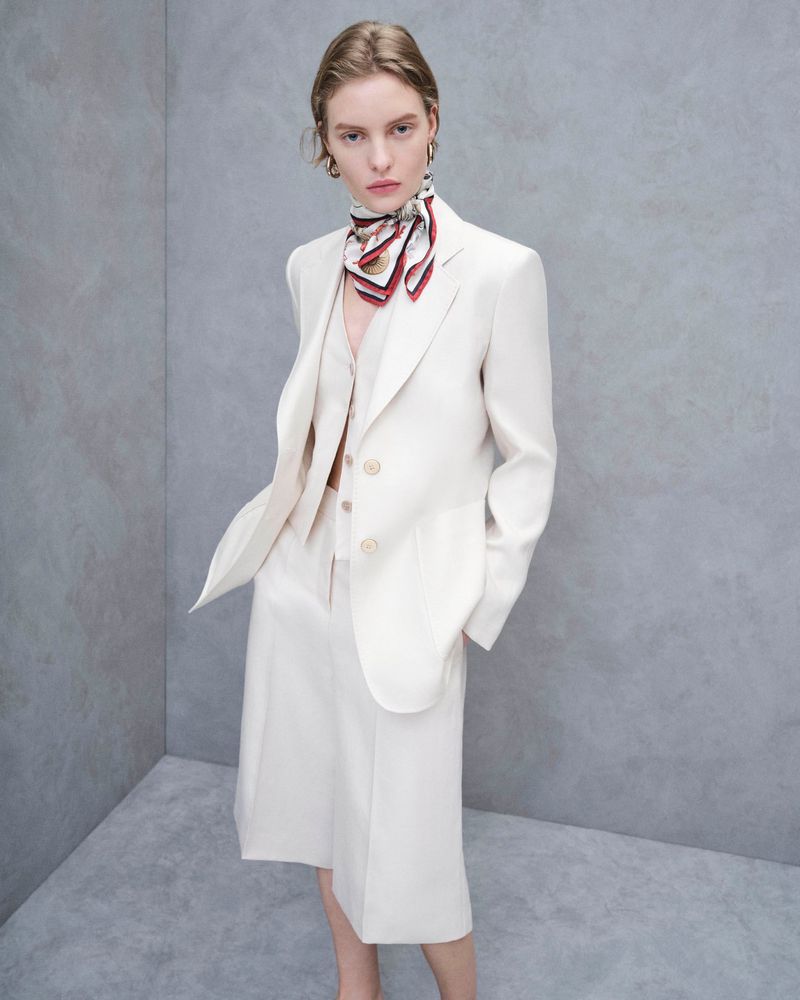 Woman in a cream-colored three-piece suit with a scarf poses against a grey backdrop in a studio shot; fashion, style, portrait.