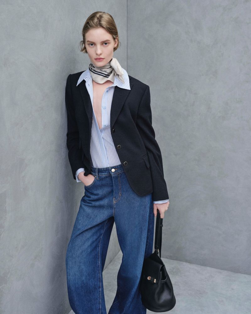 Woman in blue jeans, striped shirt, and black blazer stands indoors against gray wall, holding black purse, minimalist fashion look.