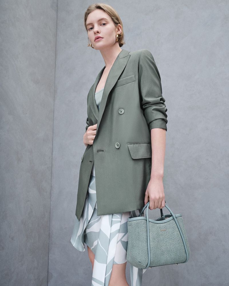 Fashionable woman in olive green blazer and patterned dress holding small handbag, posing in a photo studio with neutral gray backdrop.