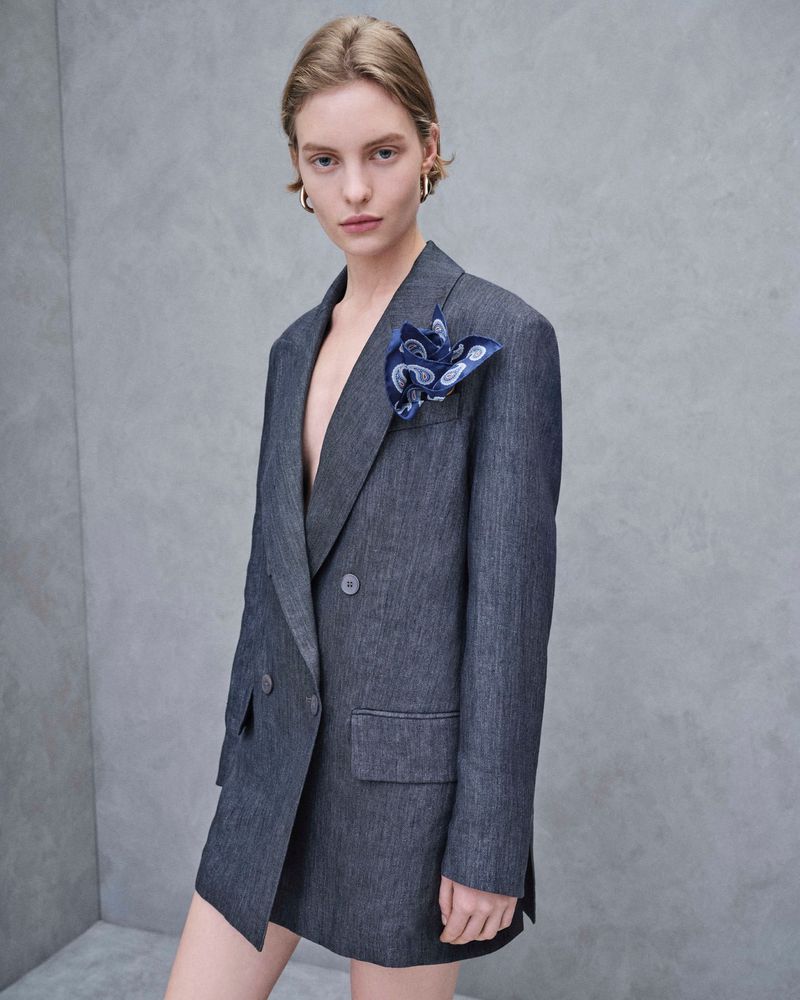 Elegant woman wearing a double-breasted blue-gray blazer dress with a patterned pocket square, posing against a neutral gray background.