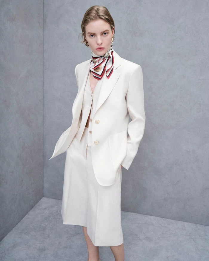 Model in a cream blazer and matching skirt wearing a patterned neck scarf against a neutral grey studio wall background