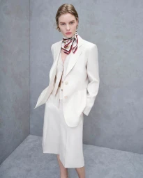 Model in a cream blazer and matching skirt wearing a patterned neck scarf against a neutral grey studio wall background