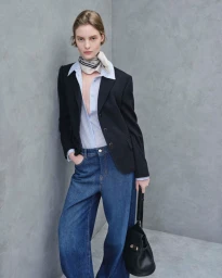 Model wearing a black blazer, striped shirt, and denim jeans standing against a textured grey wall