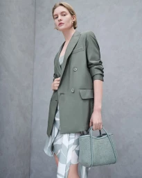 Model in olive green blazer and patterned skirt holding a blue handbag against a grey textured studio backdrop