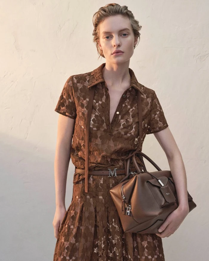Model in brown lace shirt and skirt holding a brown leather handbag against a neutral wall