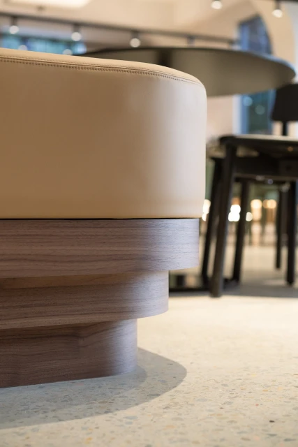 Close-up of a cushioned stool with wooden base, standing on a terrazzo floor, with black chair and circular table in soft-focus background.