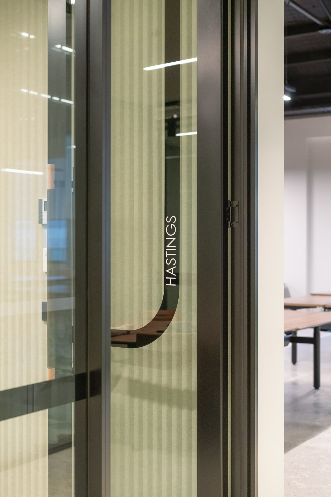Interior office space featuring a glass door with a metal frame and handle, and a room with tables and a beige wall in the background.