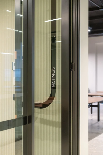 Interior office space featuring a glass door with a metal frame and handle, and a room with tables and a beige wall in the background.