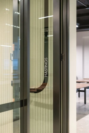 Interior office space featuring a glass door with a metal frame and handle, and a room with tables and a beige wall in the background.
