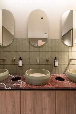 Contemporary bathroom interior with three oval mirrors, stone sinks, and brushed gold fixtures against a sage green tiled wall.