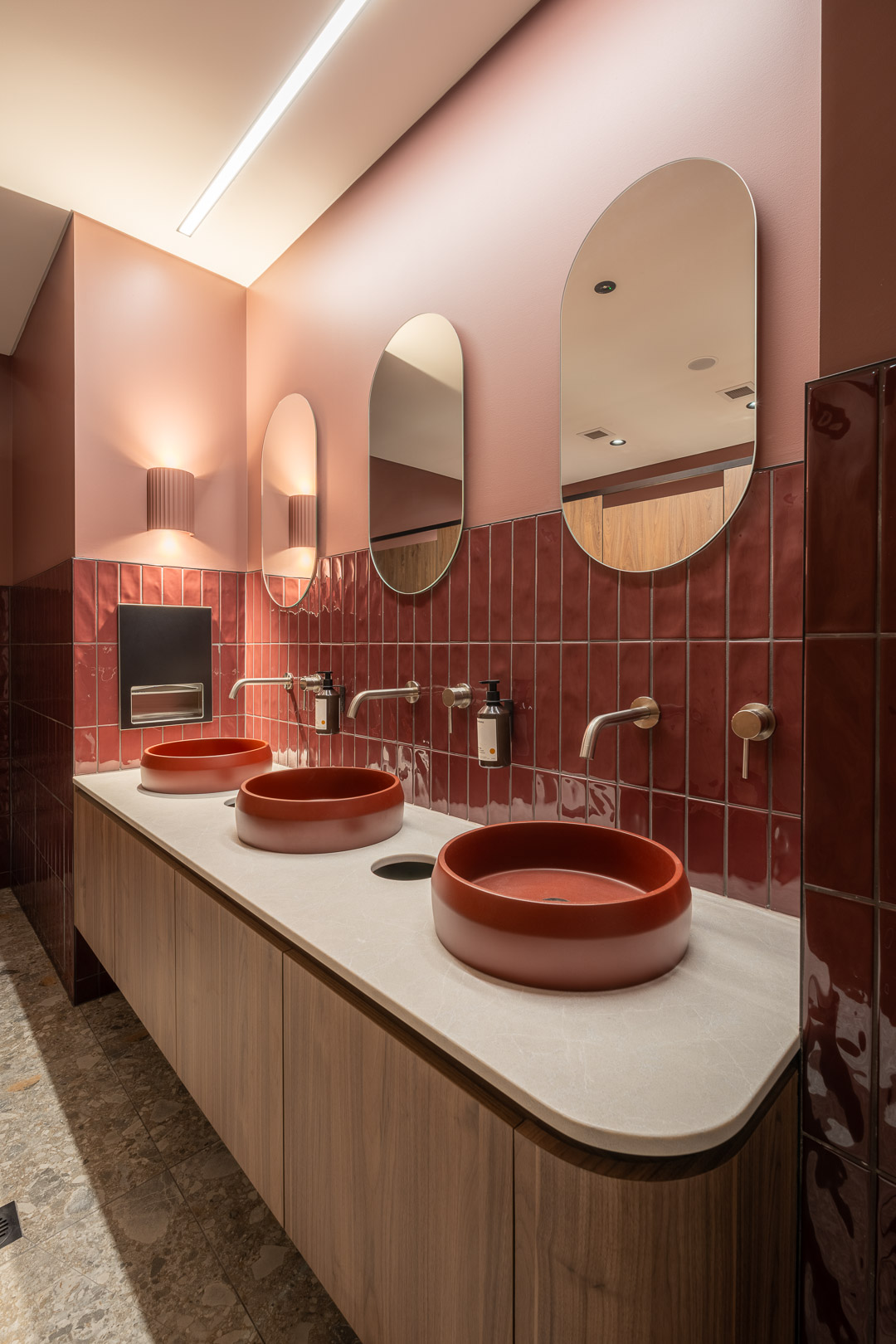 Modern bathroom interior with three red vessel sinks on a wooden vanity, set against a red tiled wall and oval mirrors.