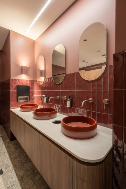 Modern bathroom interior with three red vessel sinks on a wooden vanity, set against a red tiled wall and oval mirrors.