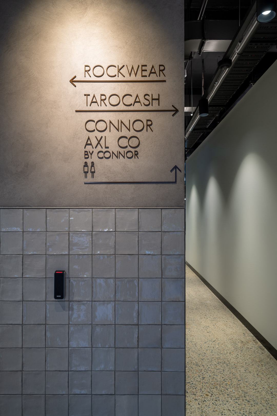Interior signage on a grey tiled wall points visitors to Rockwear, Tarocash and Connor/AXL Co in a modern building.