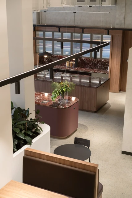 Indoor cafe with tables, chairs, booth seating, and a marble-topped serving counter with decorative plants in a modern setting.