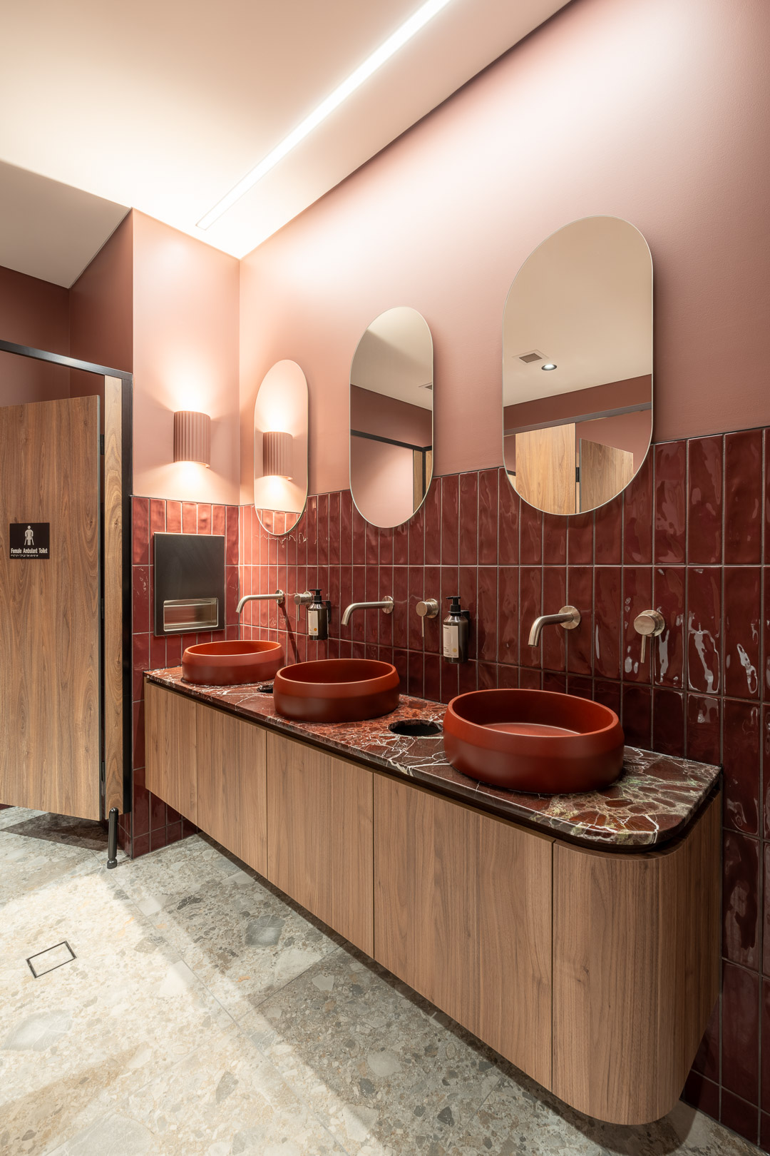 Modern public restroom interior featuring three red vessel sinks, wood cabinetry, oval mirrors, and decorative lighting fixtures.