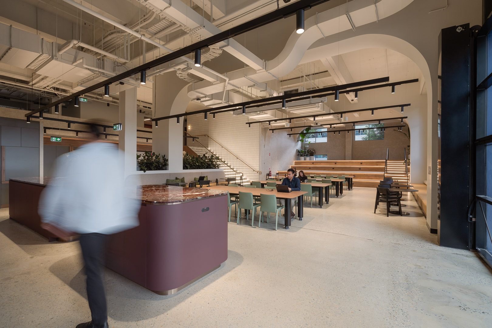 Spacious office interior features tables, chairs, stairs, and large windows. People work at tables under track lighting in a modern workspace.