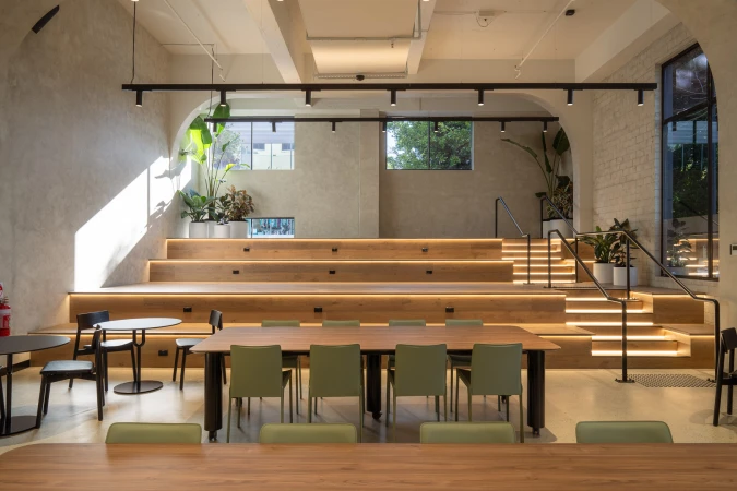 Interior view of a modern office space with wooden tiered seating, a large table with chairs, and plants in white pots, creating a bright workspace.