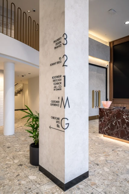 A tall column in a building interior displays floor directory information with black lettering and numerals, alongside potted plant and desk.