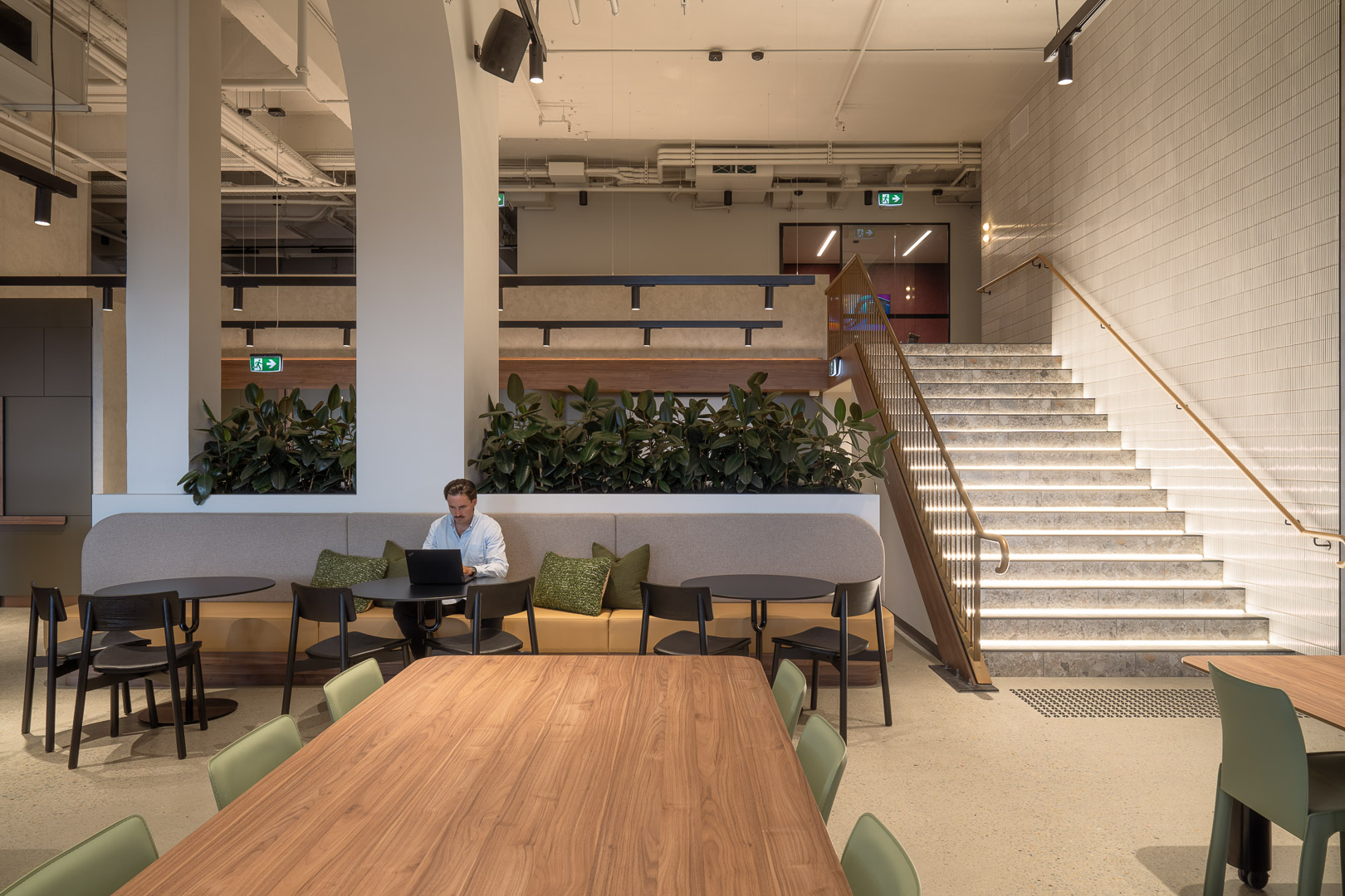 Open office space with a man working on a laptop at a table, a staircase with embedded lights, and potted plants along a bench.