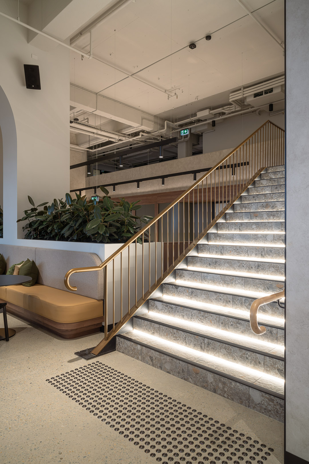 Interior view of a modern staircase with golden railing and illuminated steps, alongside a built-in beige bench with green plants.