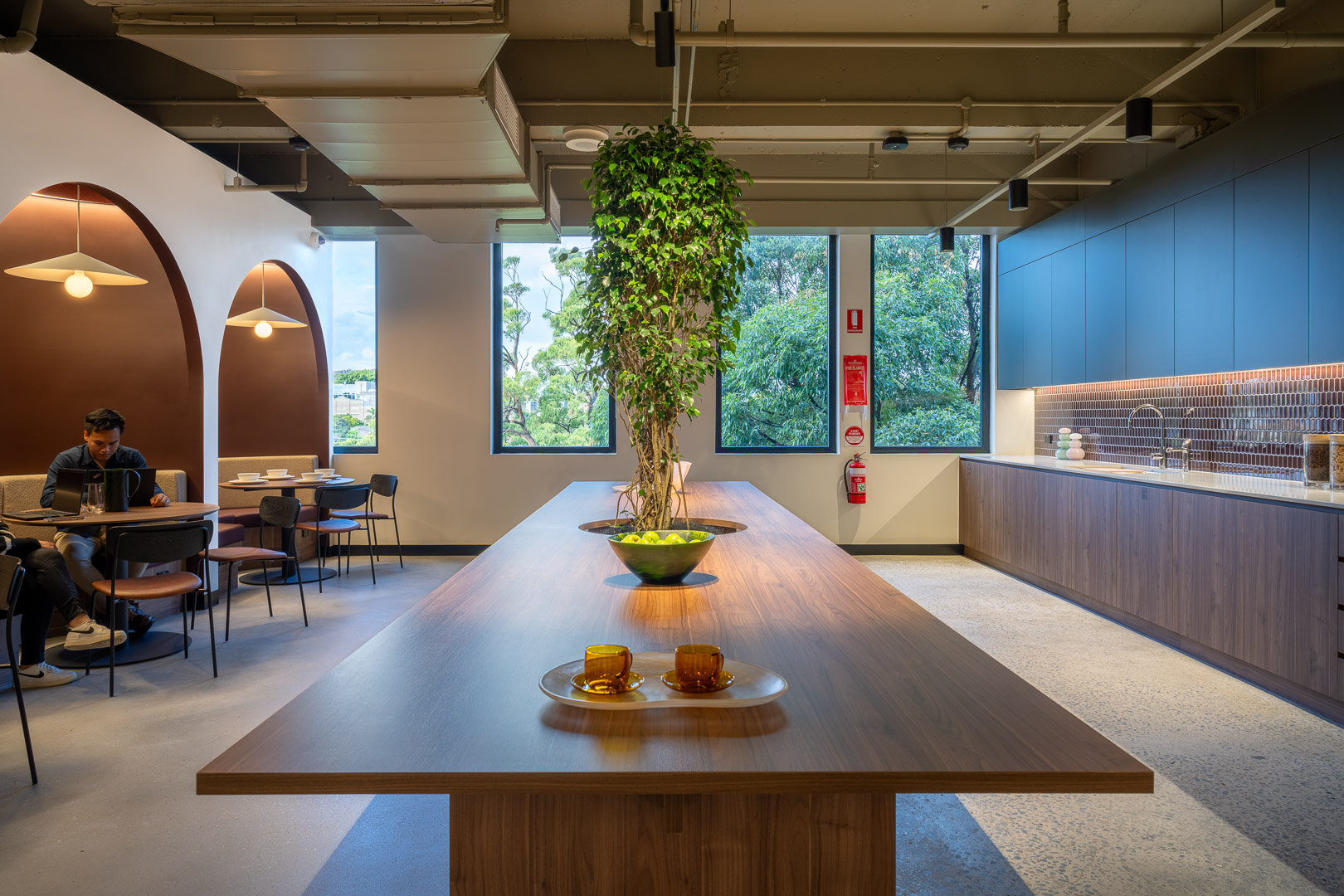 Office interior featuring long wooden table with decorative plant, surrounded by seating areas and a kitchenette, creating a modern workspace.