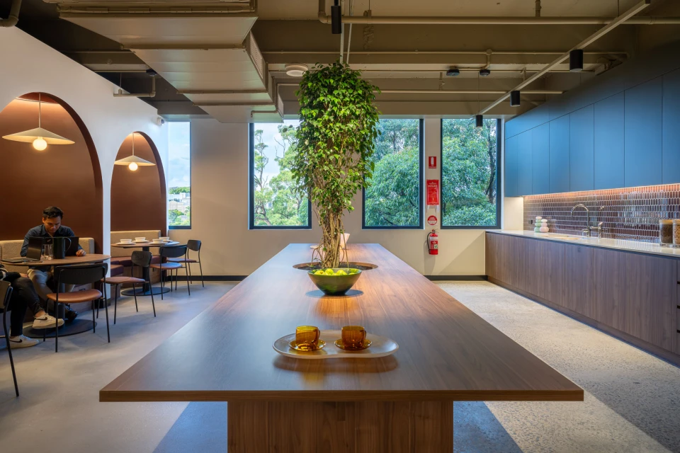Office interior featuring long wooden table with decorative plant, surrounded by seating areas and a kitchenette, creating a modern workspace.