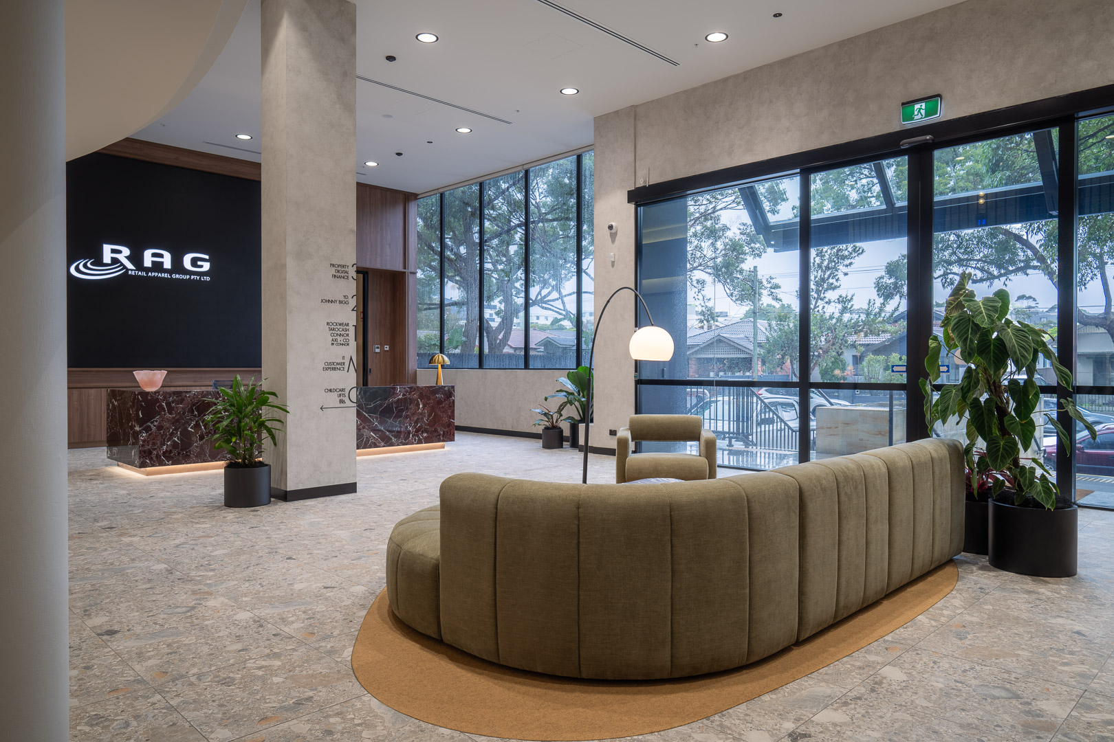 Bright, modern lobby with a rounded couch, armchairs, potted plants, marble reception desks, and large windows overlooking the outdoors.
