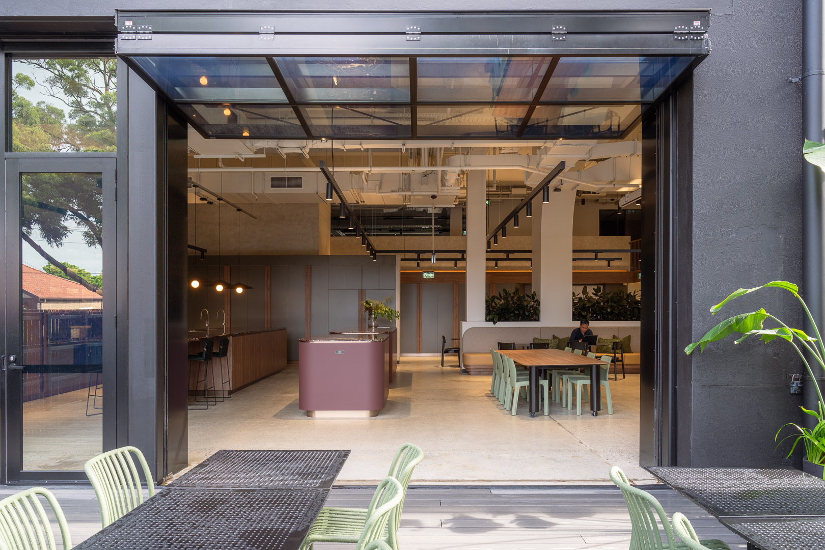 Interior of modern office space with open floor plan, featuring tables, chairs, and kitchen area, visible through open black metal doors.