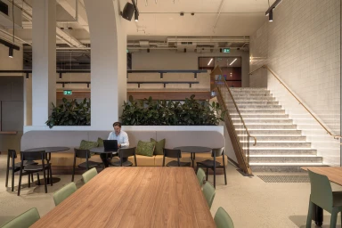 Man seated at a table working on a laptop in a modern commercial office interior with a lit staircase