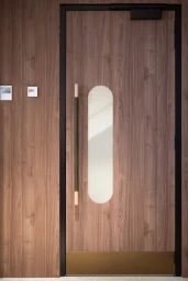 Modern wooden door with a vertical glass cutout panel and a dark handle set against matching wood panels.
