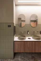 Modern commercial restroom featuring dual oval vessel sinks, rounded vertical mirrors, and sage green wall tiles by Intermain
