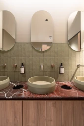 Modern commercial bathroom vanity with red marble countertop, dual green vessel sinks, and three arched capsule mirrors.