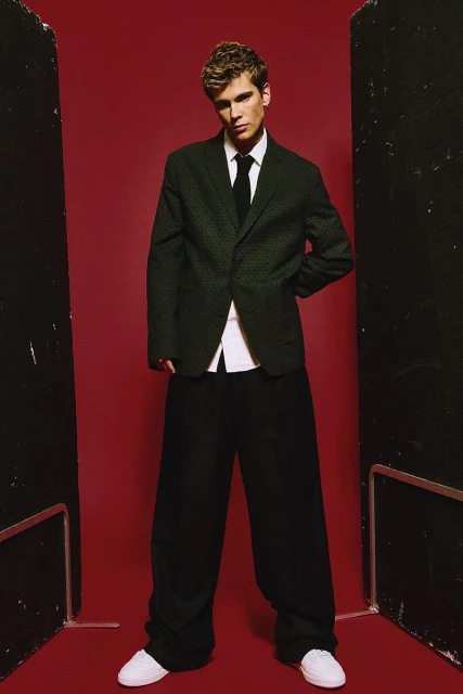 A young adult male posing indoors wearing a dark green suit, black tie, oversized black pants, and white shoes, against a dark red backdrop.