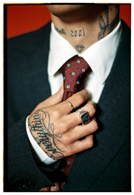 Close-up of a tattooed adult male in a suit adjusting a patterned tie, against a red background, emphasizing fashion and personal style.