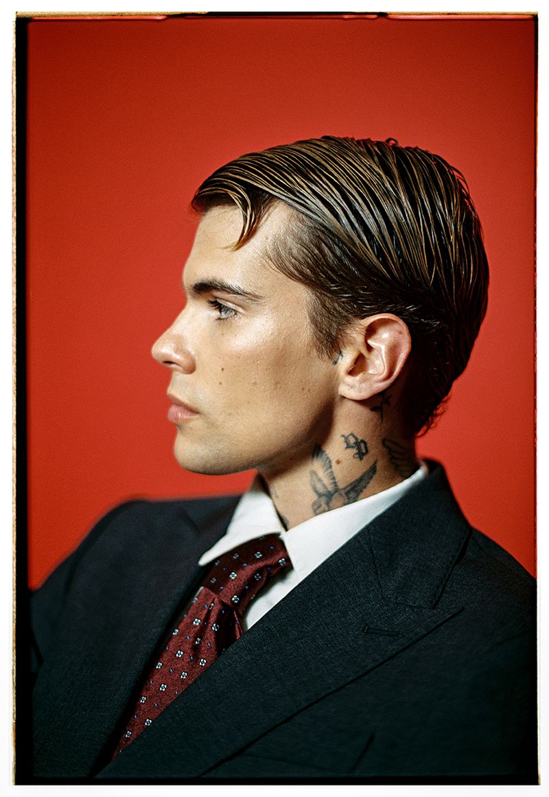 Profile portrait of a handsome adult male with a neck tattoo wearing a suit and tie against a red backdrop in a photo studio.