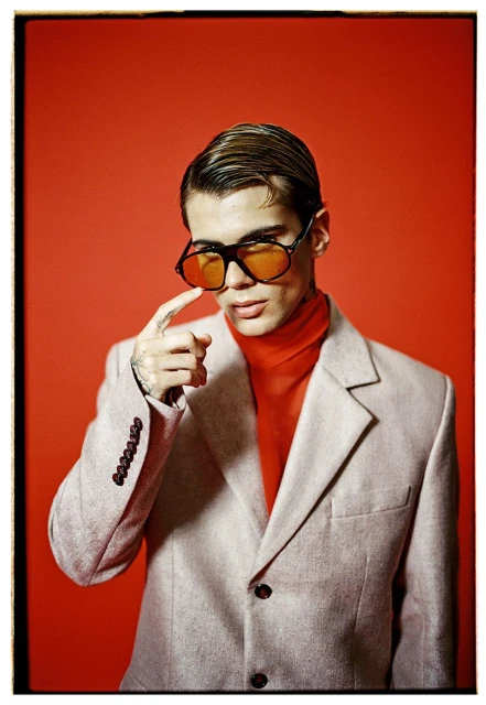 Close-up portrait of a young adult male in a gray blazer and orange turtleneck wearing large, tinted glasses against a red background.